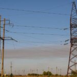 Two large utility towers standing in a field with a blue sky behind them and several smaller utility poles in the distance.