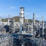 A sprawling modern industrial petroleum refining plant with several hills and a blue sky in the background.