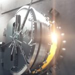 Off-center view of a large, circular bank vault door opening. Golden light shines out of the doorway toward the viewer.