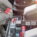 A gloved hand holding a red-plastic-coated car fuse between the thumb and index finger over a car's engine.