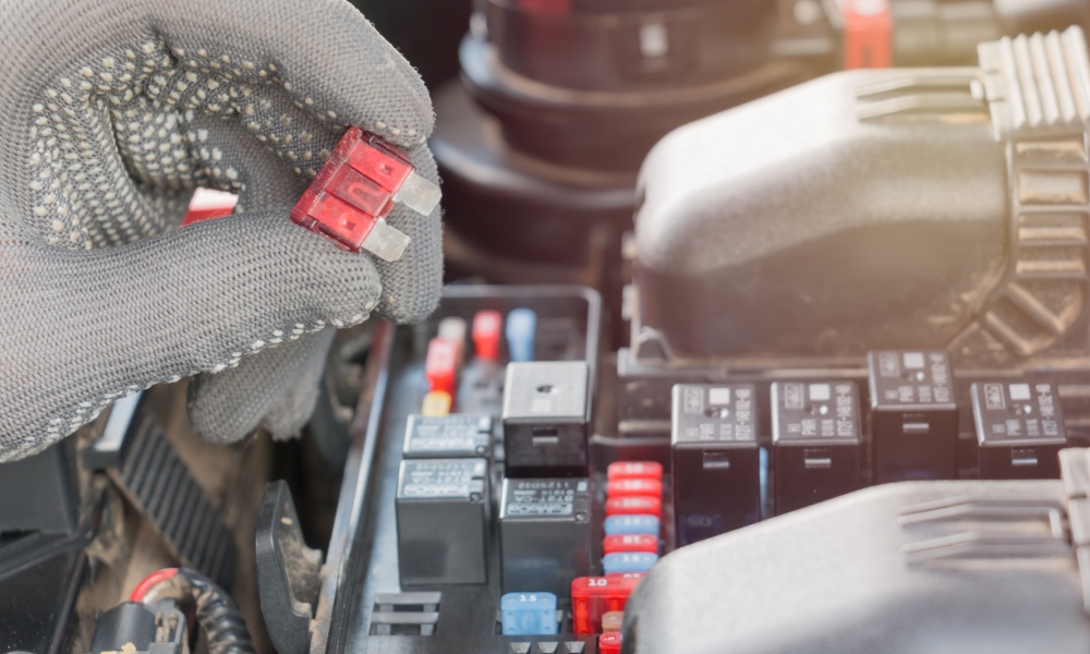 A gloved hand holding a red-plastic-coated car fuse between the thumb and index finger over a car's engine.