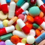 Many pills of various colors lie together, including green, blue, and white products. They are either capsules or tablets.