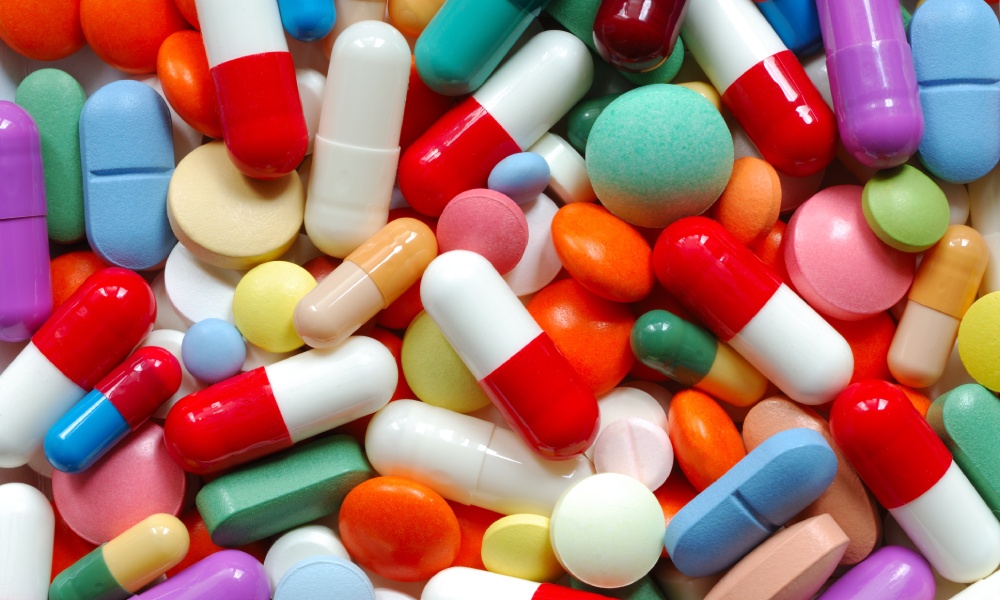 Many pills of various colors lie together, including green, blue, and white products. They are either capsules or tablets.