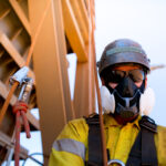 Male industrial rope access welder wearing P3 mask, mandatory protection harness, and helmet safety equipment.