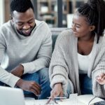 Couple of young adults smiling at each other while sitting on their living room couch looking at a laptop, files, and a calculator.