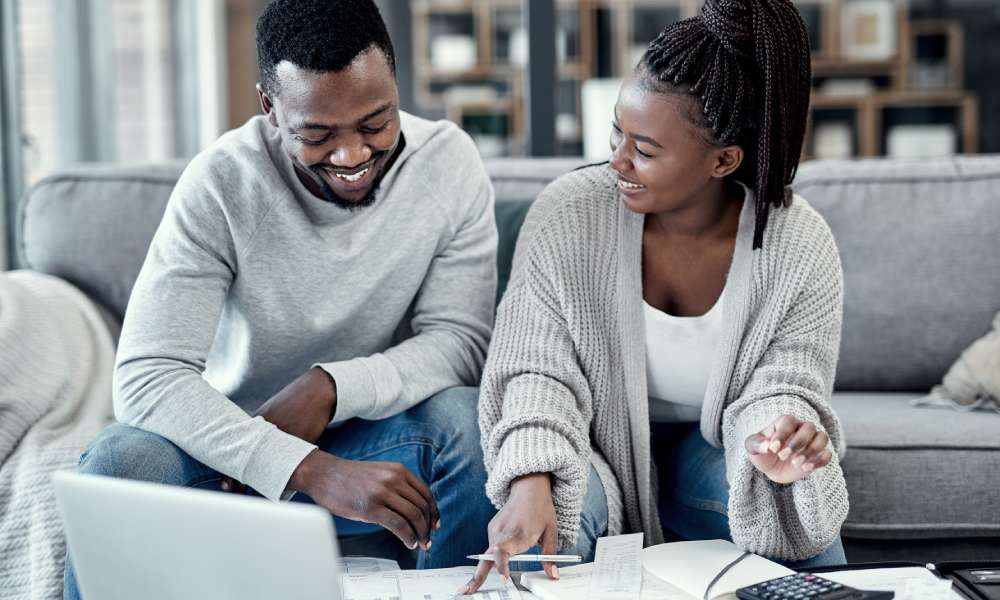 Couple of young adults smiling at each other while sitting on their living room couch looking at a laptop, files, and a calculator.