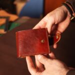 A person’s hands holding a small brown leather pouch with a snap closure. Leatherworking supplies sit on a nearby table.
