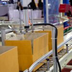 A production line’s metal conveyor belt carrying three cardboard boxes beneath a series of packing mechanisms.