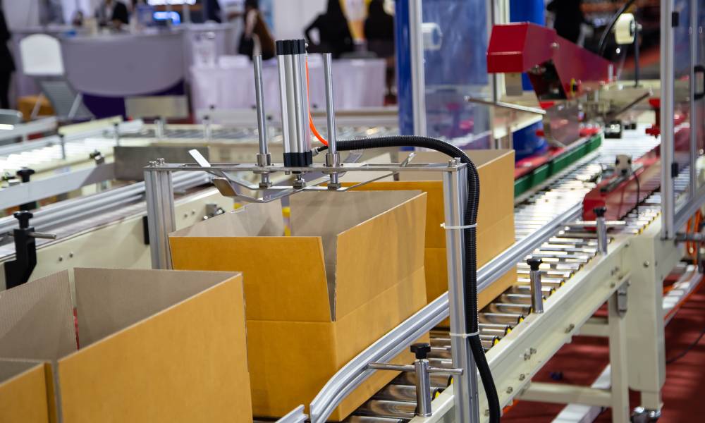 A production line’s metal conveyor belt carrying three cardboard boxes beneath a series of packing mechanisms.