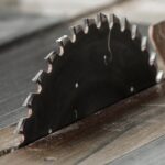 A close-up view of a circular saw cutting a piece of metal. The saw has toothed edges and there is dust on the metal.