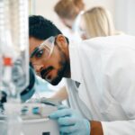 A lab worker looks at a piece of lab equipment. He wears safety glasses, a white coat, and blue gloves.