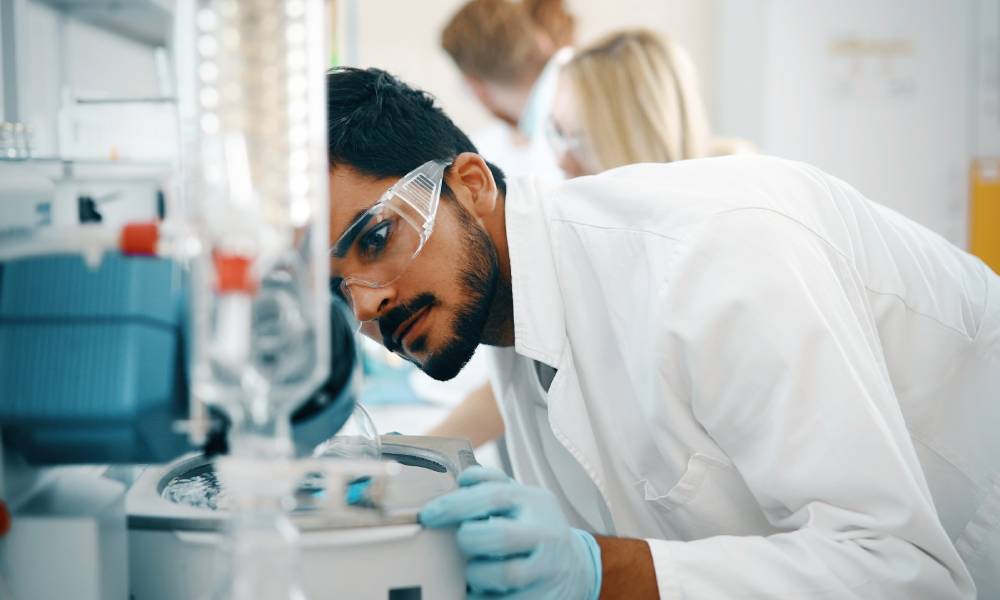 A lab worker looks at a piece of lab equipment. He wears safety glasses, a white coat, and blue gloves.