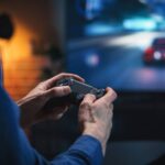 A gamer in a blue sweater holding out a black controller as they make progress in the racing game visible on the TV.