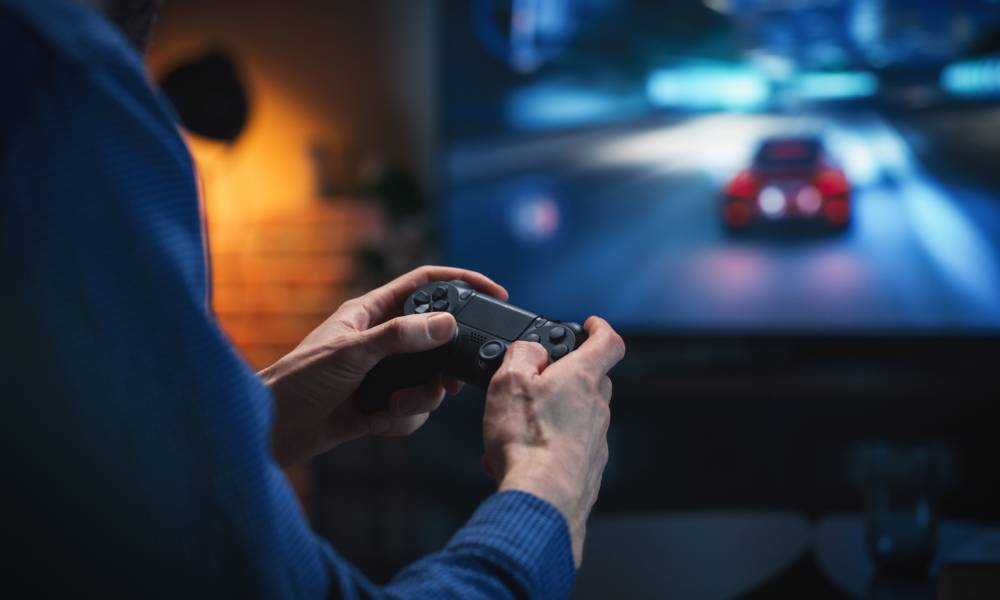 A gamer in a blue sweater holding out a black controller as they make progress in the racing game visible on the TV.