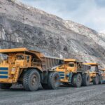 Five yellow mining vehicles sit behind each other in a line at a large open-air mining facility. The sky is cloudy and gray.