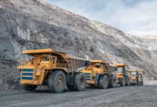 Five yellow mining vehicles sit behind each other in a line at a large open-air mining facility. The sky is cloudy and gray.