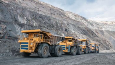 Five yellow mining vehicles sit behind each other in a line at a large open-air mining facility. The sky is cloudy and gray.