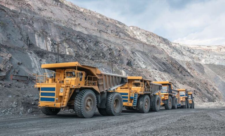 Five yellow mining vehicles sit behind each other in a line at a large open-air mining facility. The sky is cloudy and gray.