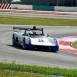 A silver race car bearing blue stripes, black spoilers, and the number 38 drives around a bend in a race track.