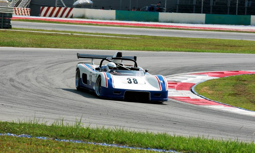 A silver race car bearing blue stripes, black spoilers, and the number 38 drives around a bend in a race track.