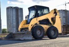 A skid steer sitting in a parking lot. There are some tall buildings in the background, and one of them is being worked on.