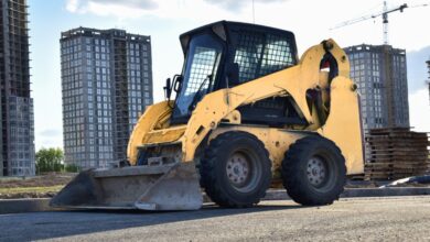 A skid steer sitting in a parking lot. There are some tall buildings in the background, and one of them is being worked on.