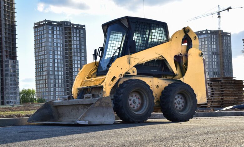 A skid steer sitting in a parking lot. There are some tall buildings in the background, and one of them is being worked on.