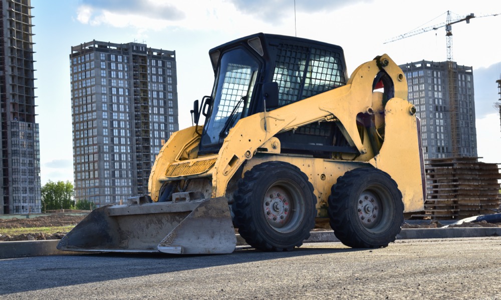 A skid steer sitting in a parking lot. There are some tall buildings in the background, and one of them is being worked on.