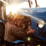 A farmer mechanic checks the engine of his blue tractor at sunset as part of his preventative maintenance schedule.