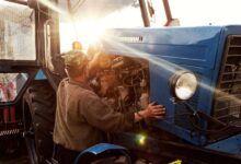 A farmer mechanic checks the engine of his blue tractor at sunset as part of his preventative maintenance schedule.