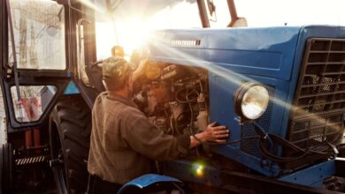 A farmer mechanic checks the engine of his blue tractor at sunset as part of his preventative maintenance schedule.