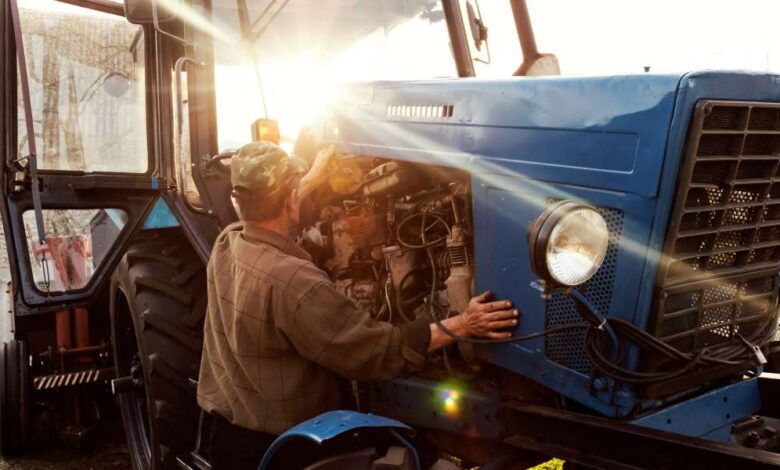 A farmer mechanic checks the engine of his blue tractor at sunset as part of his preventative maintenance schedule.