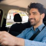 A man with wavy dark hair and a button-down blue shirt sits behind the wheel of a vehicle. He is smiling.
