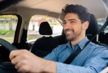 A man with wavy dark hair and a button-down blue shirt sits behind the wheel of a vehicle. He is smiling.