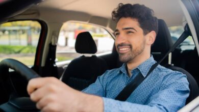 A man with wavy dark hair and a button-down blue shirt sits behind the wheel of a vehicle. He is smiling.