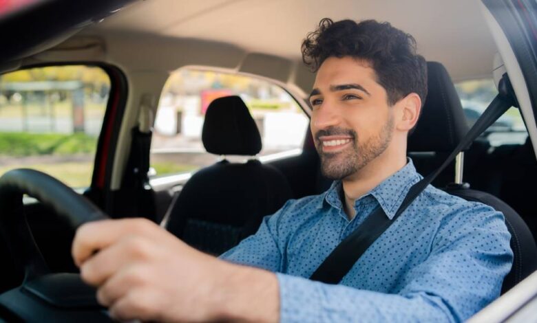 A man with wavy dark hair and a button-down blue shirt sits behind the wheel of a vehicle. He is smiling.