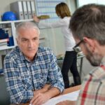 A man sitting across a desk from another man talking about information on a piece of paper inside an office environment.