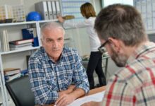 A man sitting across a desk from another man talking about information on a piece of paper inside an office environment.