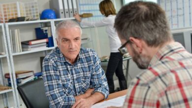 A man sitting across a desk from another man talking about information on a piece of paper inside an office environment.