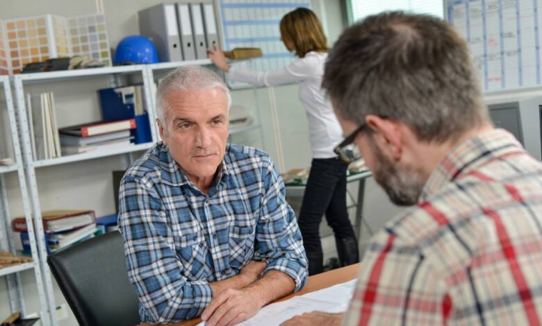 A man sitting across a desk from another man talking about information on a piece of paper inside an office environment.