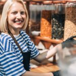 A woman smiles as she wears an apron and gloves and works in a health food store. She stands in front of containers of nuts.