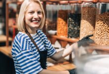 A woman smiles as she wears an apron and gloves and works in a health food store. She stands in front of containers of nuts.