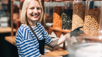 A woman smiles as she wears an apron and gloves and works in a health food store. She stands in front of containers of nuts.