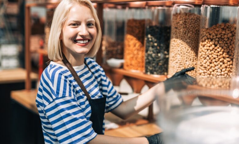 A woman smiles as she wears an apron and gloves and works in a health food store. She stands in front of containers of nuts.