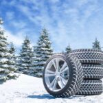 A set of four winter tires with three stacked on top of one another resting on a snowy hill with snowy trees behind them.