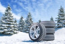 A set of four winter tires with three stacked on top of one another resting on a snowy hill with snowy trees behind them.