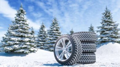 A set of four winter tires with three stacked on top of one another resting on a snowy hill with snowy trees behind them.