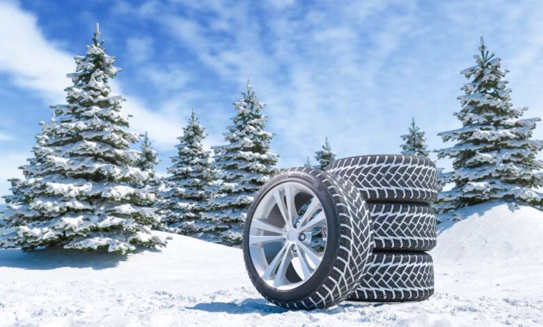 A set of four winter tires with three stacked on top of one another resting on a snowy hill with snowy trees behind them.