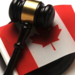 Close-up of a brown and gold gavel sitting on top of a small Canadian flag. The gavel is on a white table.