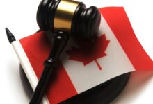 Close-up of a brown and gold gavel sitting on top of a small Canadian flag. The gavel is on a white table.